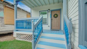 Un porche con barandillas azules y una puerta de madera, que da a una casa con revestimiento blanco.