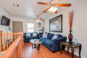 A living room with a blue sofa, a glass coffee table, a wooden side table, a ceiling fan, and a mounted TV.