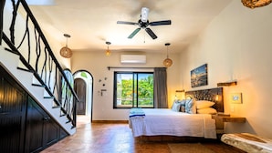 A bedroom with a bed, a ceiling fan, a window with curtains, a wooden bench, and a staircase with black railings.