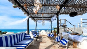 Un área de descanso frente a la playa con asientos a rayas azul y blanco, una mesa de madera con bebidas y vista al océano.