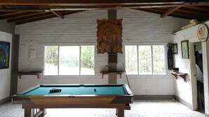 A pool table in a room with white brick walls, wooden beams on the ceiling, and windows with a view of trees.