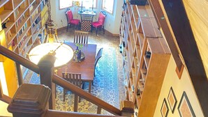 Una stanza con soffitto alto, una scala in legno, un'area pranzo con un lampadario e vista su un cielo azzurro attraverso ampie finestre.