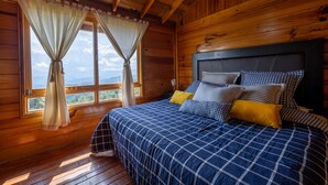 A wooden cabin bedroom with a bed, curtains, and a view of the outdoors. - Cabin in Private Forest (MORELIA)
