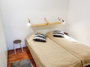 Una habitación pequeña con cama individual, suelo de madera, mesita de noche, lámpara y estante en la pared.