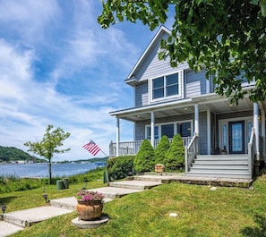 A two-story house with a porch, a flag, and a view of the water. - Open year round for adventure and relaxation.  Cozy winter escape less than 20 minutes from Crystal Mountain! (Frankfort)