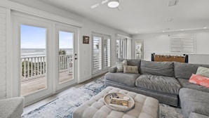 A spacious living room with a grey sofa, a wooden headboard, and a view of the ocean through large sliding doors.