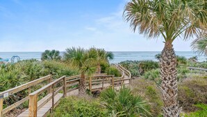 A wooden boardwalk leading to a beach with palm trees and coastal vegetation.