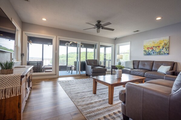 A spacious living room with a large sofa, a coffee table, and a fireplace. There is a balcony with a view of the outdoors. - Take in Million dollar views on the screened in porch or large deck. (Camdenton)