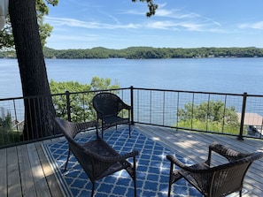 A deck with wicker chairs and a blue patterned rug, overlooking a lake with a forested shoreline.