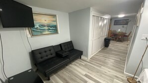Ett vardagsrum med en svart soffa, en platt-TV som är monterad på väggen, en tavla och en matplats med ett bord och stolar.