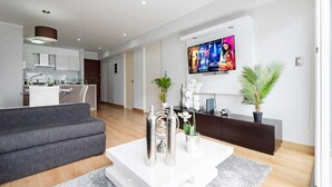 Una sala moderna con un sofá gris, una mesita de centro blanca y elementos decorativos. Un televisor está montado en la pared mostrando carteles de películas.