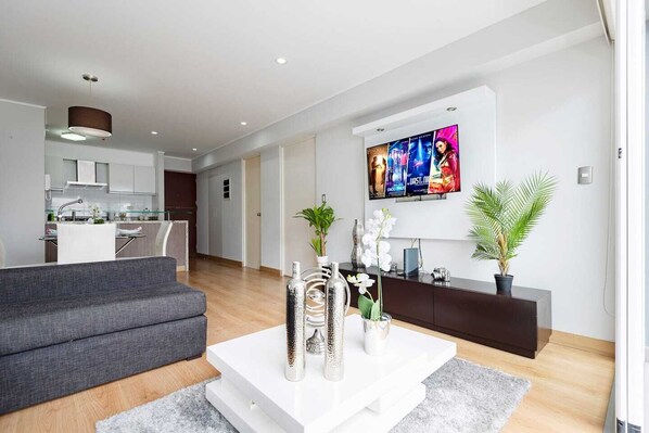 Una sala moderna con un sofá gris, una mesita de centro blanca y elementos decorativos. Un televisor está montado en la pared mostrando carteles de películas.
