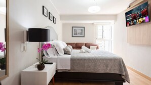 Un dormitorio con cama, mesita de noche con lámpara, televisor montado en la pared y ventana con vistas.