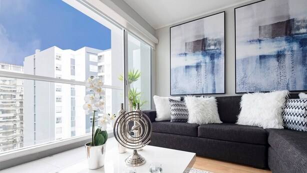 Una sala de estar moderna con un sofá gris, cojines blancos y estampados, una mesa de centro con objetos decorativos y grandes cuadros abstractos en la pared.