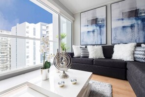 Una sala de estar moderna con un sofá gris, cojines blancos y estampados, una mesa de centro con objetos decorativos y grandes cuadros abstractos en la pared.