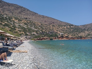 Una playa de guijarros con agua cristalina, sombrillas y gente tomando el sol.