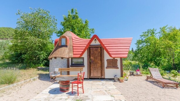 A small, rustic house with a red roof and stone walls, a wooden bench, a table, and a chair in the foreground, and a lounge chair in the background. - Immaculate 2-Bed Cottage near Krka Waterfalls (Rupe)