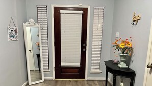 A hallway with a wooden floor, a dark brown door with white blinds, a mirror, and a small table with a vase of flowers. - 4110 David & Jennie’s Place (Missouri City)