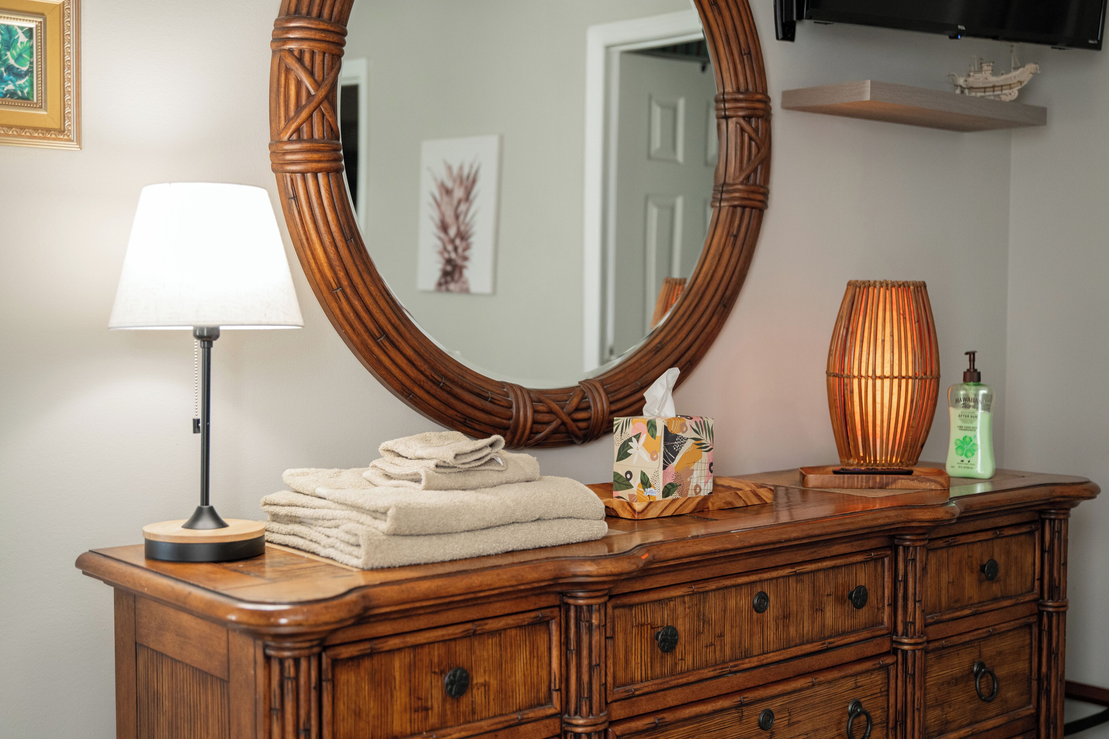 A wooden dresser with a large mirror, a lamp, folded towels, a decorative basket, and a bottle of hand soap.