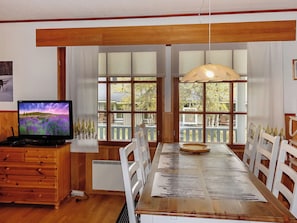 Ein Essbereich mit Holztisch, weißen Stühlen, einem Fernseher auf einer Holzkommode und großen Fenstern mit Blick auf Bäume und einen Zaun.