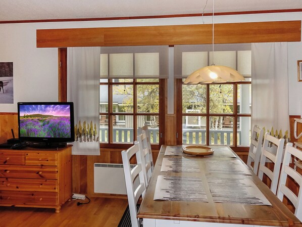 Ein Essbereich mit Holztisch, weißen Stühlen, einem Fernseher auf einer Holzkommode und großen Fenstern mit Blick auf Bäume und einen Zaun.