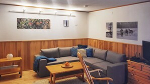 Una sala de estar acogedora con un sofá gris, una mesa de centro de madera y un sillón mecededor. Hay una mesita auxiliar con un frutero y una lámpara. La habitación tiene paneles de madera en las paredes y una lámpara en el techo.