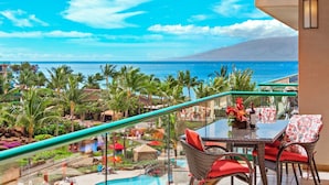A balcony with a glass railing overlooking a pool and a tropical resort area with palm trees and a view of the ocean.
