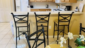 A kitchen with wooden cabinets, a white refrigerator, a microwave, and a sink. There are three black chairs with a woven seat and backrest.
