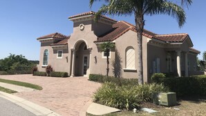 A two-story house with a tiled roof, a central entrance with an archway, and a small garden area with palm trees and shrubs.