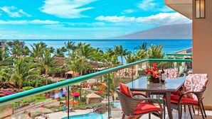 A balcony with a glass railing overlooking a pool and a tropical resort area with palm trees and a view of the ocean.