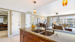 A modern kitchen with granite countertops, stainless steel sinks, and wooden cabinets. There is a dining area with a table and chairs, and a balcony with a view of the city.