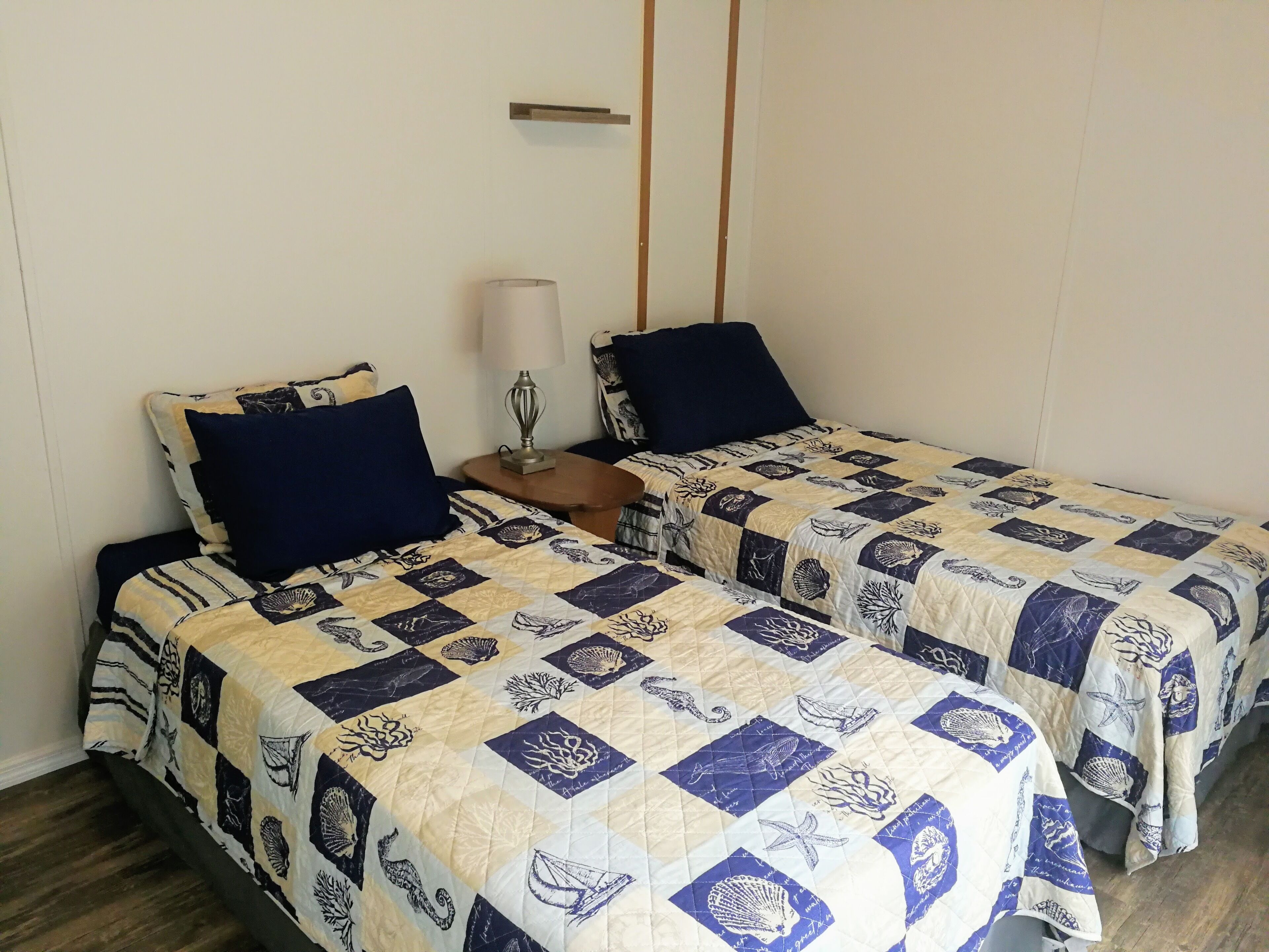 Une chambre avec deux lits, chacun doté d’une tête de lit matelassée et d’un oreiller. Une petite table avec une lampe se trouve entre les deux lits.