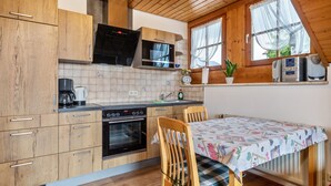 Een keuken met houten kastjes, een magnetron, een oven, een tafel met een tafelkleed met bloemenmotief, en een raam met een plant.