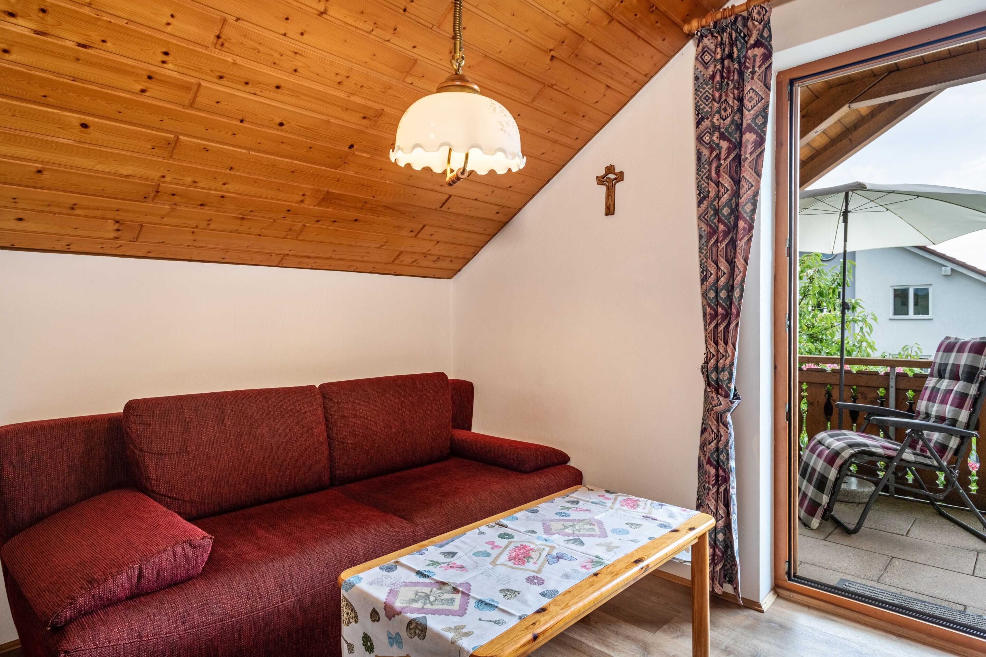 Un plafond en bois, un canapé rouge, une petite table recouverte d’une nappe à motifs et une vue sur un patio extérieur avec un fauteuil.