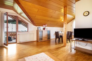 Amplia sala de estar con piso de madera, un televisor grande sobre un mueble, comedor con sillas y una puerta corrediza de vidrio que da a un área exterior.