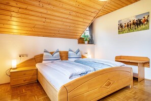 Un dormitorio con techo de madera, una cama con ropa de cama a rayas, una mesita de noche, un buró y una repisa en la pared con una planta, además de un cuadro de vacas.