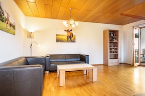 Un salón con techo de madera, un sofá de cuero negro, una mesa de centro de madera, una estantería y un cuadro de una vaca en la pared.