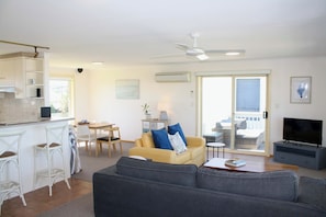 A living room with a sofa, a television, and a dining area with chairs and a table. - Esplanade Escape, Beachfront, Whale Watching, Surf (Middleton)