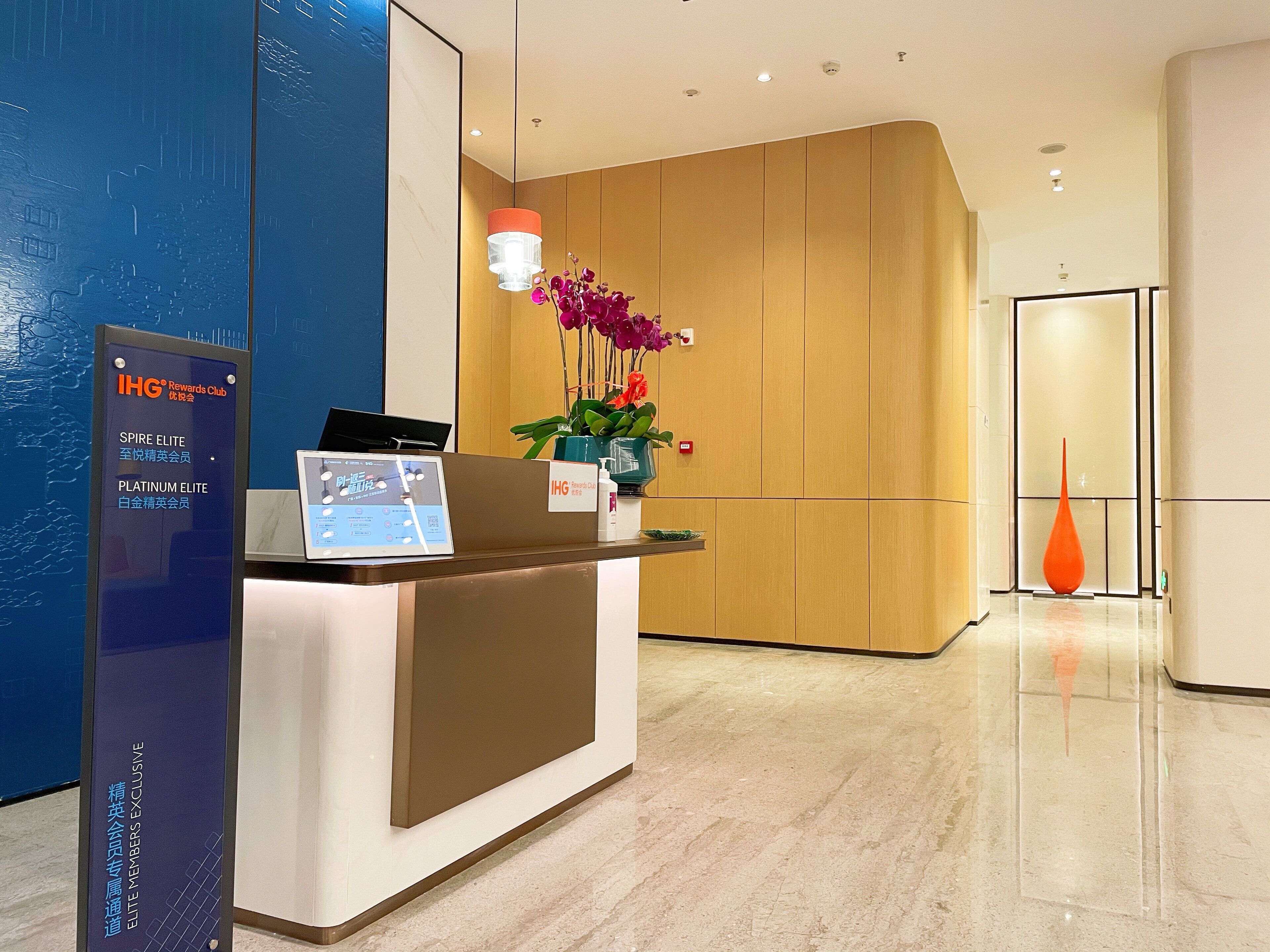 A hotel reception area with a modern design, featuring a reception desk, a digital screen, and a decorative orchid arrangement.