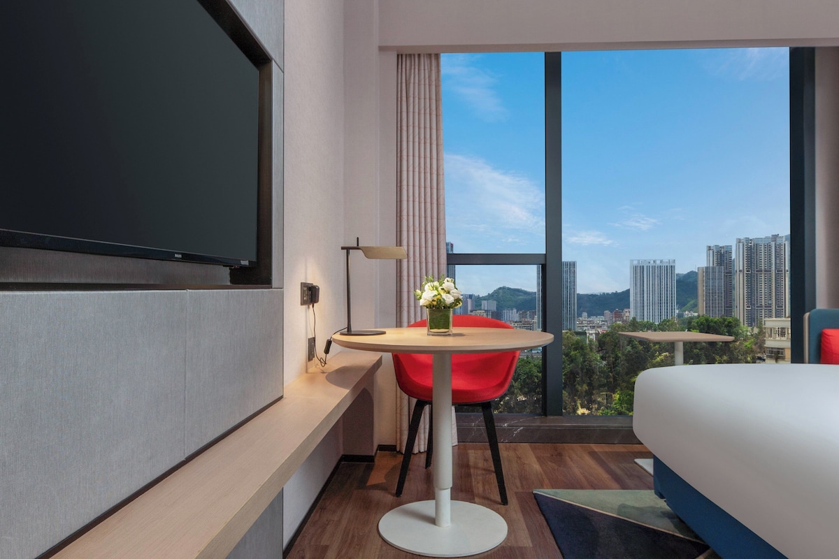 Hotelkamer met een bed, een aan de muur bevestigde televisie, een ronde tafel met een rode stoel en een groot raam met uitzicht op de stad.