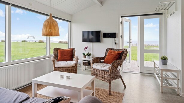 A bright room with wicker chairs, a white table, and a TV. Large windows offer a view of the outdoors. - Charming 2-bedroom 12H Ocean Retreat, Summerhouse (Middelfart)