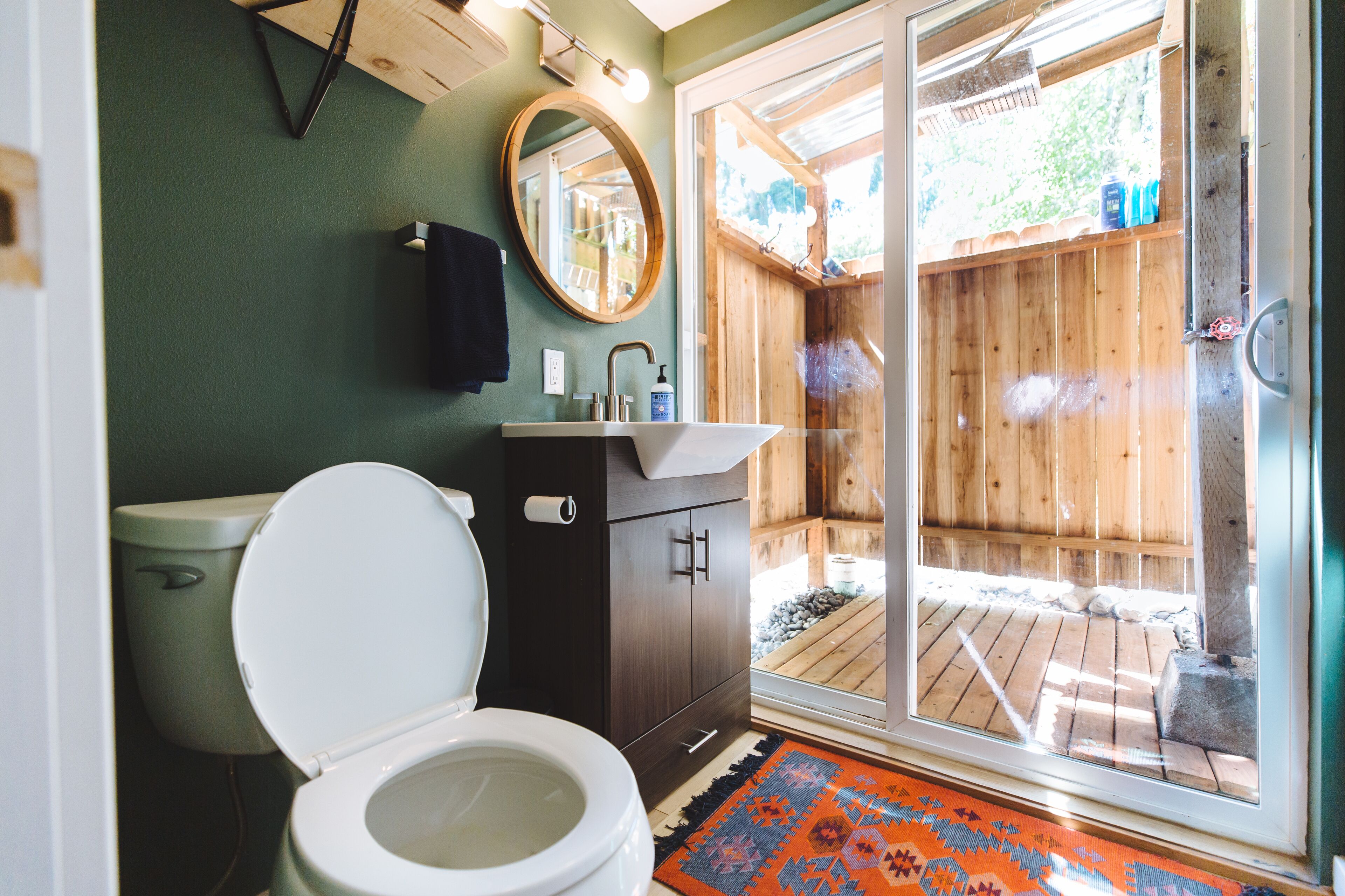 Baño con inodoro, lavamanos, espejo y una terraza de madera visible a través de una puerta de cristal.