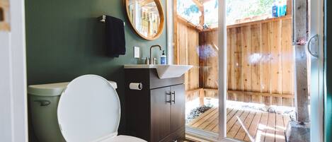Baño con inodoro, lavamanos, espejo y una terraza de madera visible a través de una puerta de cristal.