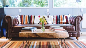 Un canapé en cuir marron, recouvert d’un plaid rayé aux couleurs vives, une table basse en bois avec des livres et un vase de fleurs, et un tapis rayé posé sur un plancher en bois.