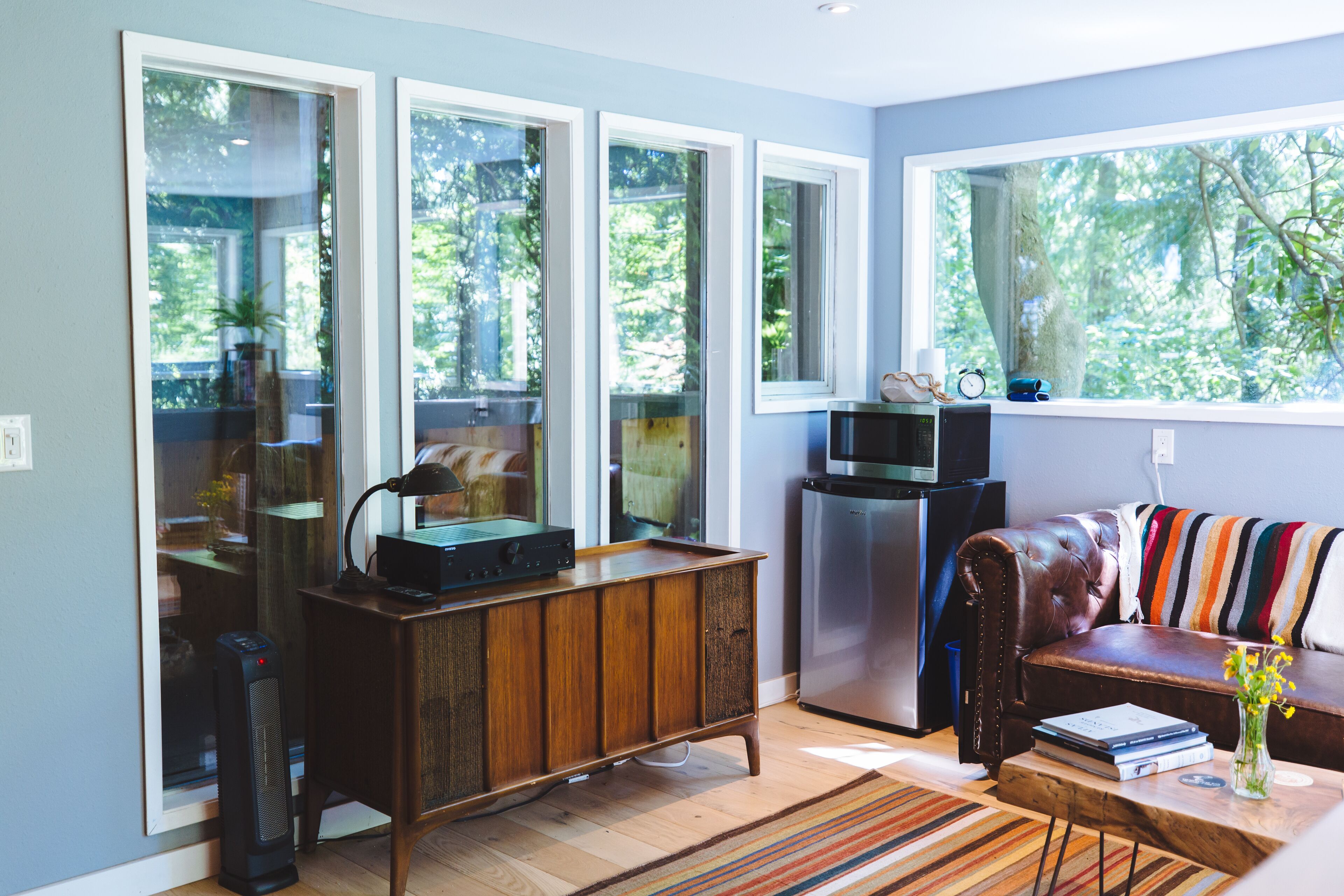 A modern living room with large windows, a leather sofa, a wooden cabinet, and a small kitchen area.