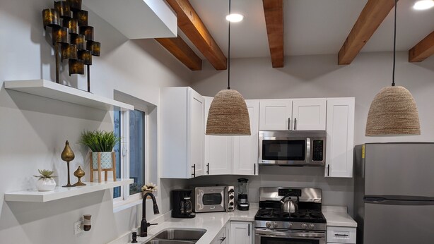 A modern kitchen with white cabinets, stainless steel appliances, a built-in oven, a microwave, and a refrigerator. There are pendant lights hanging from the ceiling and a window with a view of the outside. - Brand New Studio, Confortable, and gated parking, close to downtown. (Ensenada)