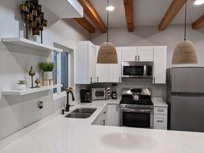 A modern kitchen with white cabinets, stainless steel appliances, a built-in oven, a microwave, and a refrigerator. There are pendant lights hanging from the ceiling and a window with a view of the outside. - Brand New Studio, Confortable, and gated parking, close to downtown. (Ensenada)