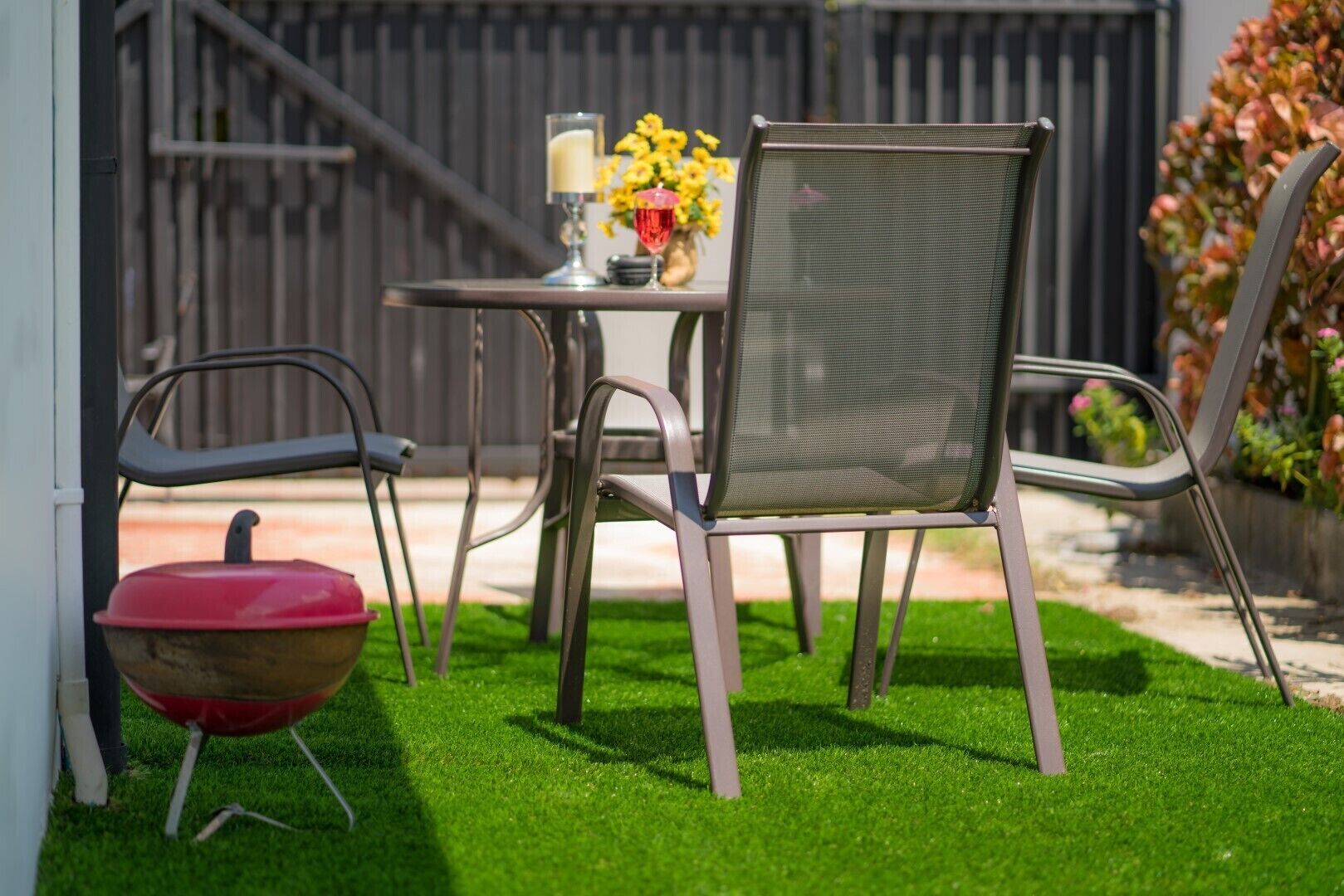 Een terras met een ronde tafel, twee stoelen, een barbecue en een vaas met bloemen.