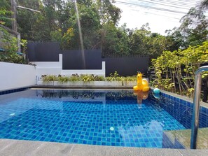 Una piscina con piso azulado de baldosas, rodeada de follaje y un muro blanco con un panel negro.