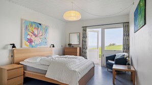 A bedroom with a bed, bedside tables, a desk, a chair, a hanging light fixture, and a large window with curtains. - Cozy Brenderup Apartment on the Beach (Middelfart)
